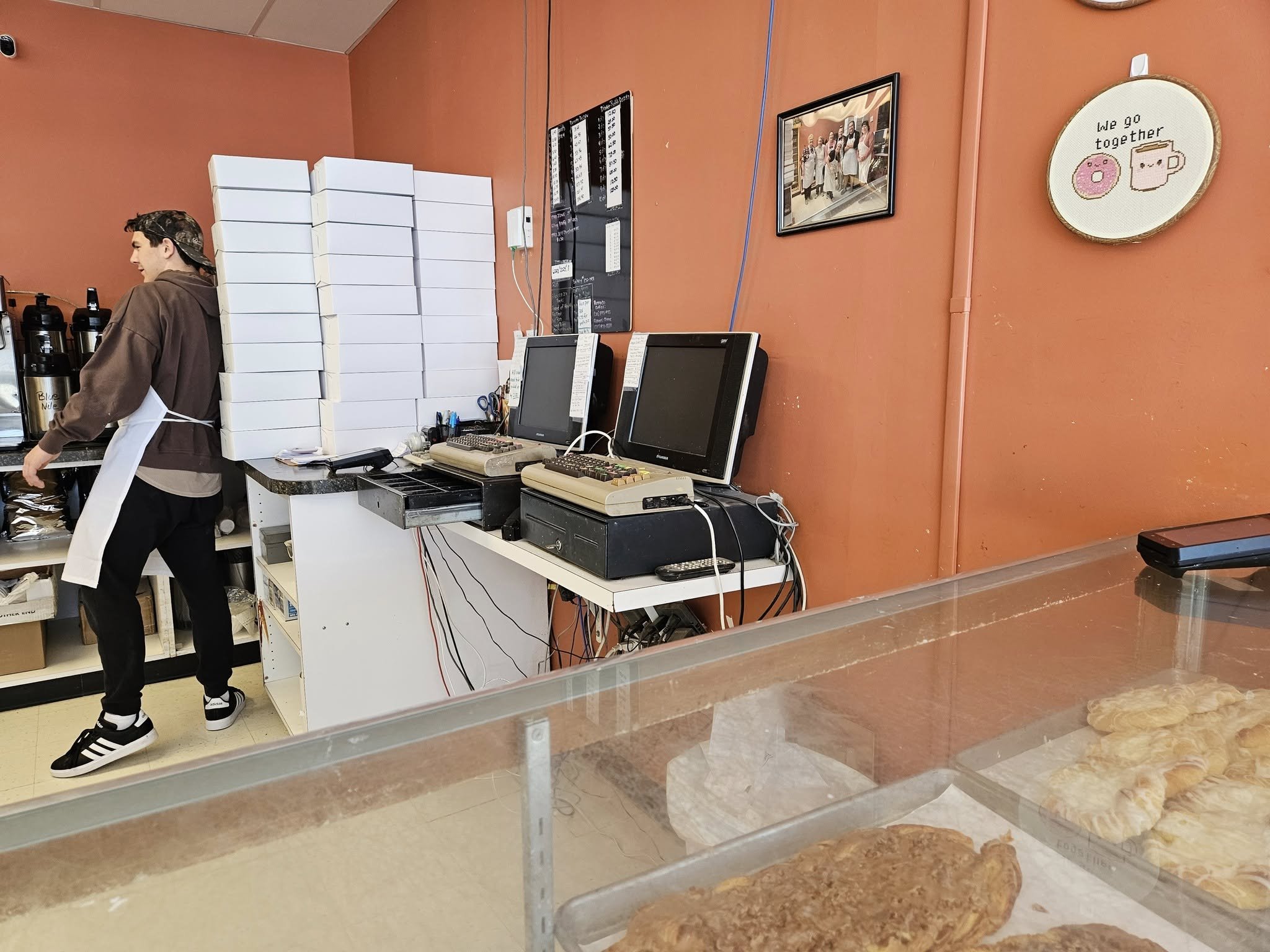 Balcão de padaria com dois computadores antigos de teclado bege e monitores de tubo, caixas de papelão brancas empilhadas e um funcionário ao fundo, atrás de v…