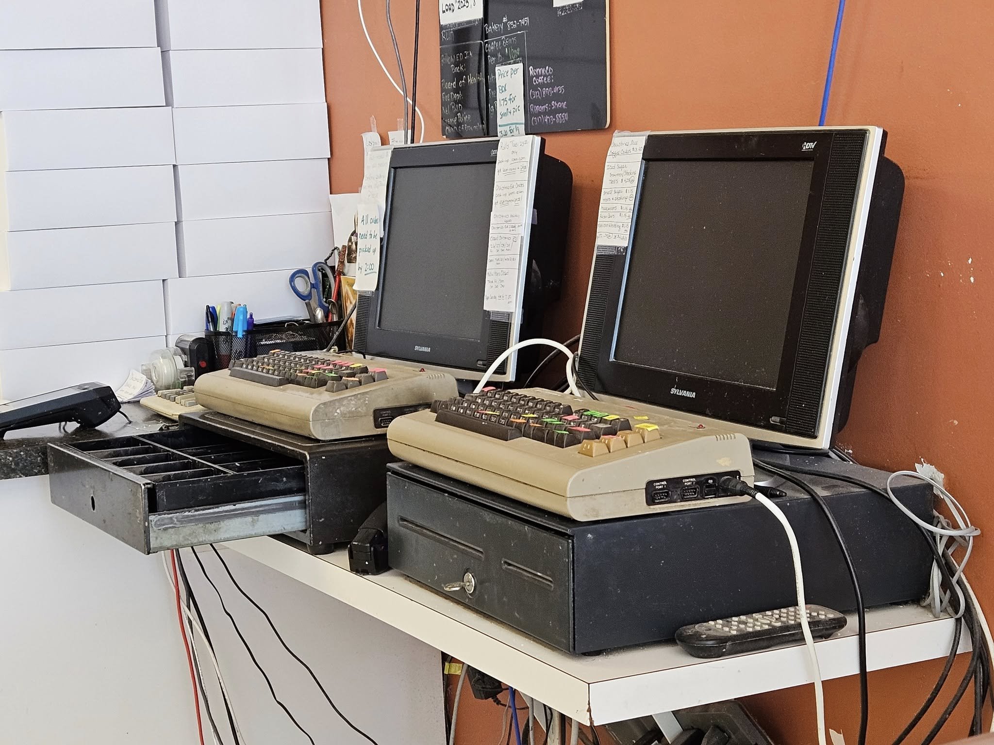Dois caixas registradores antigos, de teclado bege com teclas coloridas, sobre gavetas de dinheiro pretas, ao lado de monitores de tubo com notas de papel cola…