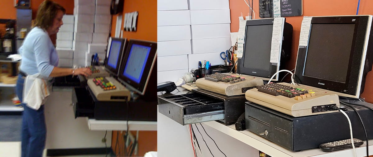Duas caixas registradoras antigas com teclados bege estilo Commodore 64 e monitores de tubo, ao lado de gavetas de dinheiro, com uma mulher digitando em uma de…