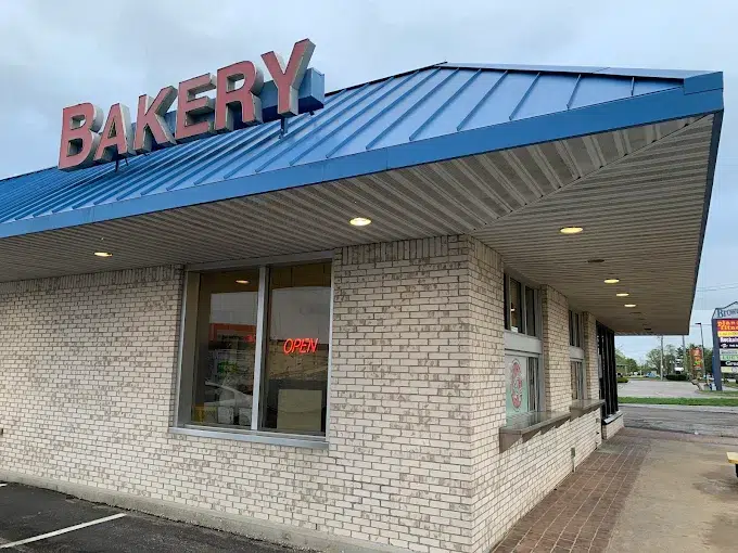 Fachada de padaria com tijolos claros, telhado metálico azul e letreiro vermelho "BAKERY", com placa de "OPEN" na vitrine
