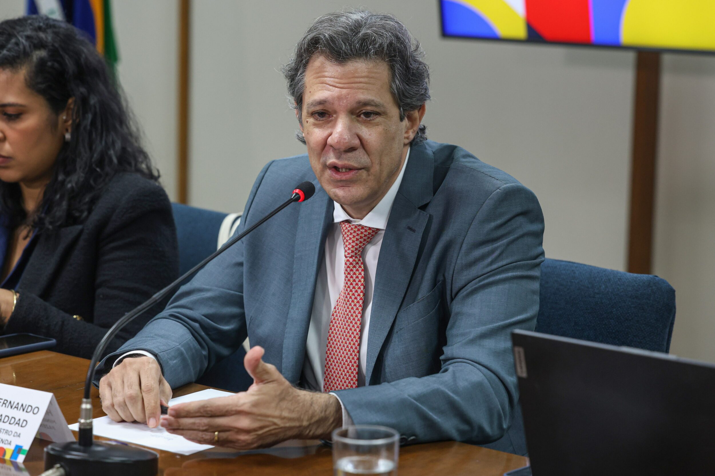 Homem de terno cinza e gravata vermelha falando em microfone de mesa, com placa de identificação "Fernando Haddad" à frente e uma mulher ao lado