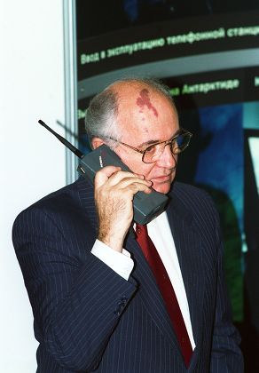 Homem idoso de terno listrado segurando um telefone celular antigo com antena no ouvido, diante de um painel com texto em russo