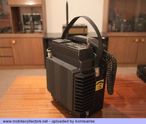 Telefone celular portátil antigo, preto, com antena e alça de transporte, apoiado sobre uma mesa de madeira em um mostruário